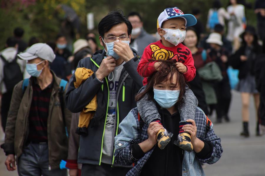 Mulher chinesa com uma criança no cangote, ambos de máscara, anda nas ruas de Pequim
