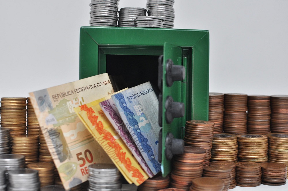 Imagem mostra notas de R$ 50, de R$ 20 e de R$ 5 saindo de um cofre entreaberto e moedas empilhadas ao redor e em cima do objeto