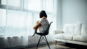 Pessoa sentada com os pés em cima da cadeira olhando para a janela, que tem uma cortina branca quase transparente. Está longe da câmera e, no canto direito, aparece metade de um sofá branco.