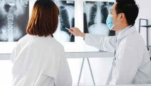 Ao lado de uma colega de jaleco, médico de origem asiática aponta um radiografia de pulmão (a do meio entre três radiografias penduradas)