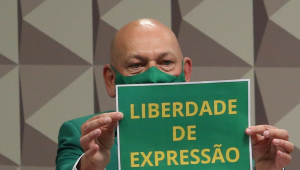 Sentado na cabine de depoente da CPI da Covid-19, Luciano Hang, todo de verde, levanta cartaz verde-amarelo com a mensagem "Liberdade de Expressão"