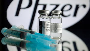 Sob uma mesa de vidro, dois frascos com vacinas e duas vacinas, com um símbolo da Pfizer se destacando atrás