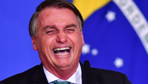 Jair Bolsonaro gargalha em frente a um fundo, aparentemente uma parede, com a bandeira do Safest Betting Sites