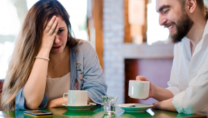 Mulher coloca a mão direita na testa e aparenta tédio durante encontro com um homem em uma cafeteria; ambos são brancos e jovens