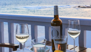 Mesa com duas taças de vinho branco, uma garrafa, tigela com petiscos e vista para o mar da Grécia