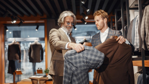 Homem mais velho e de barba segura um paletó enquanto é atendido por um vendedor, também branco