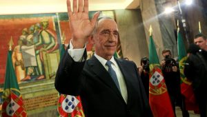 Homem branco usando terno com a mão levantada em frente a duas bandeiras de Portugal
