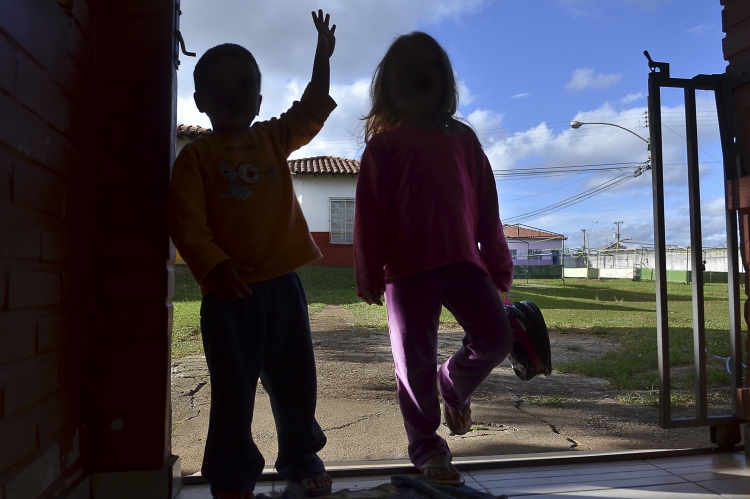 Duas crianças paradas em frente a um parque. Uma delas está com a mão acenando