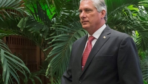 presidente de cuba, homem branco com cabelos brancos, gravata vermelha e terno preto, de pé em frente a folhas verdes