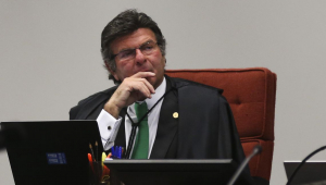 O ministro Luiz Fuz sentado em uma cadeira com estofado vermelho, usando óculos, trajes sociais e colocando a mão direita no queixo