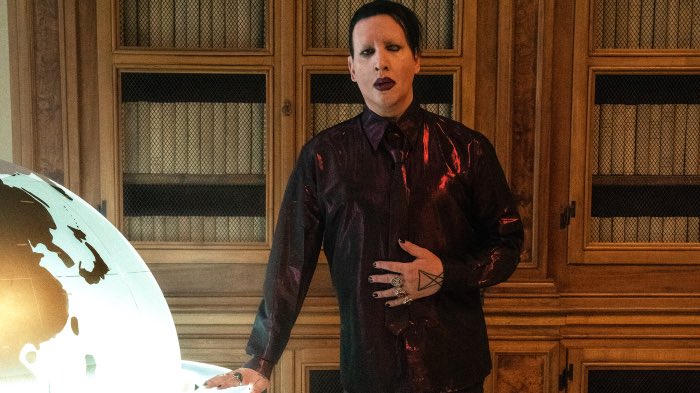Marilyn Manson com um terno de couro preto, batom e cabelos pretos, com uma das mãos em cima do terno e outra em cima da mesa. Atrás, uma estante de madeira de livros