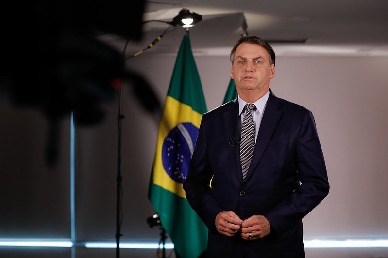 O presidente Jair Bolsonaro em pé em frente a uma bandeira do Brasil