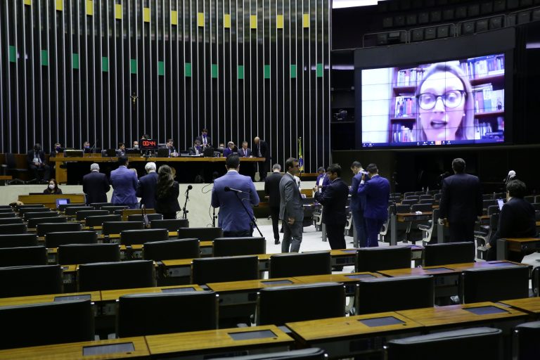 Sessão na Câmara dos Deputados, com alguns parlamentares de pé, mesas com computadores e uma deputada falando através de um telão