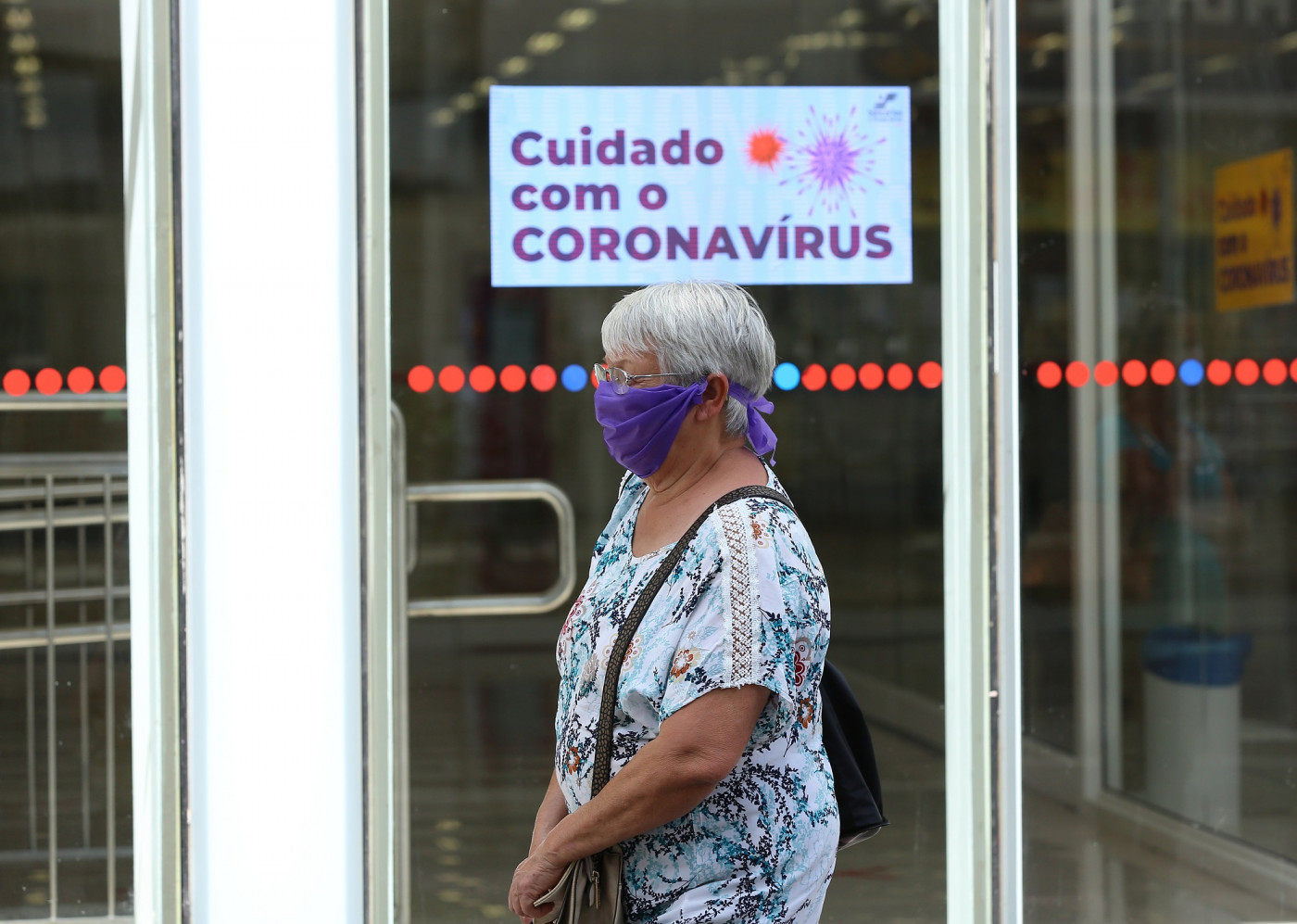 Idosa com máscara passando em frente de uma porta com a placa escrito 'Cuidado com o Coronavírus'