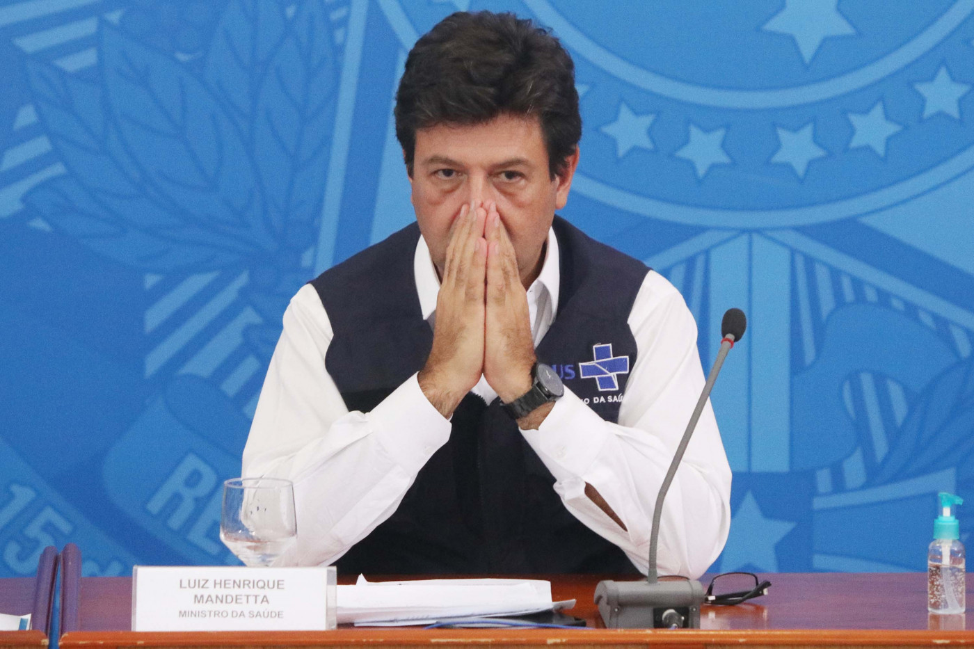 Ministro Luiz Henrique Mandetta com as mãos fechadas como se estivesse rezando. Ele usa camisa branca e um colete azul marinho do SUS. Fala em uma coletiva, está em cima de uma mesa de madeira, com um fundo azul e um microfone.
