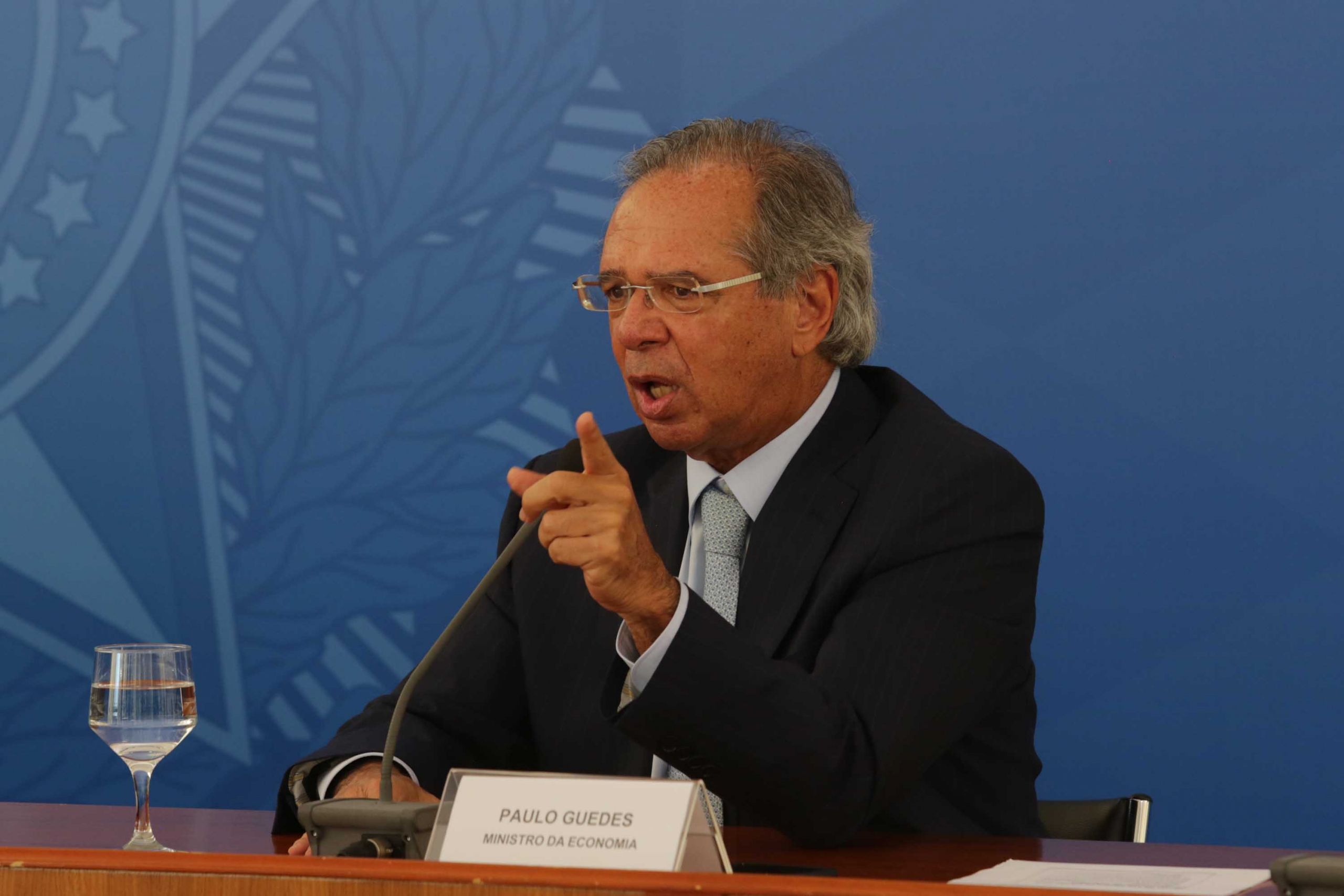 Paulo Guedes, ministro da Economia, fala com o dedo em riste, sentado à frente de um fundo azul