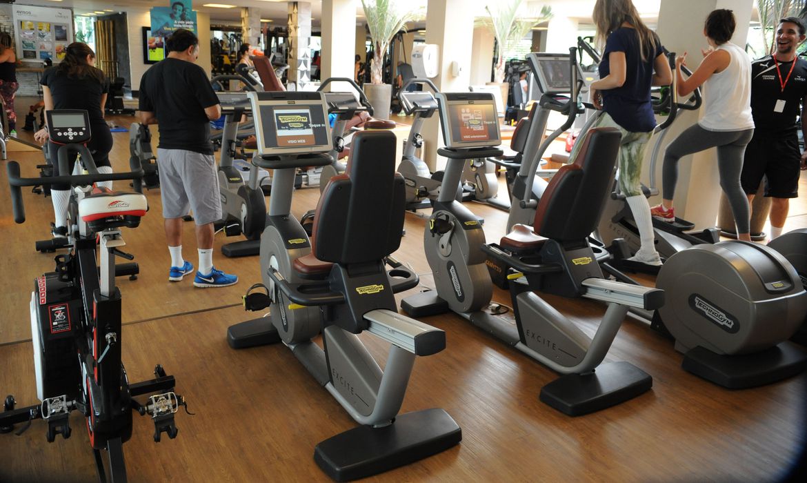 Duas máquinas de pedalar de academia colocadas sobre um chão de taco marrom em primeiro plano; no fundo, uma série de pessoas fazem exercícios físicos em outras máquinas