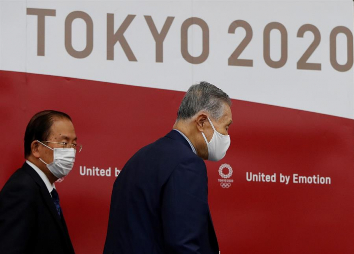 Duas pessoas caminhando de máscaras em frente a um painel escrito Tokyo 2020