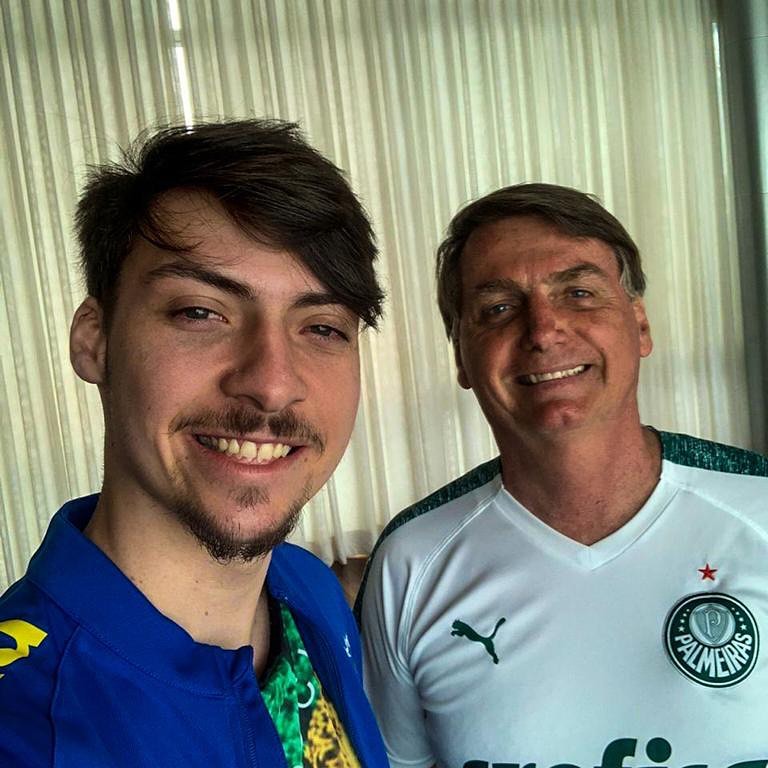 Jair Renan está ao lado do presidente Jair Bolsonaro. Os dois estão sorrindo e usam camisas de time de futebol