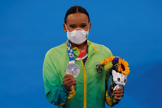 Usando máscara e o uniforme verde da delegação brasileira, Rebeca Andrade segura o mascote dos Jogos e um buquê de flores na mão esquerda e a medalha, e a medalha com a mão direita