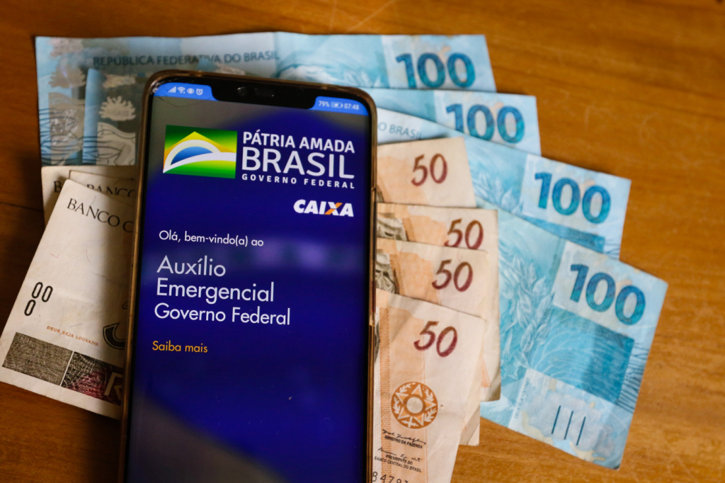Um celular logado no aplicativo da Caixa para o recebimento do auxílio emergencial e notas de dinheiro ao lado do aparelho