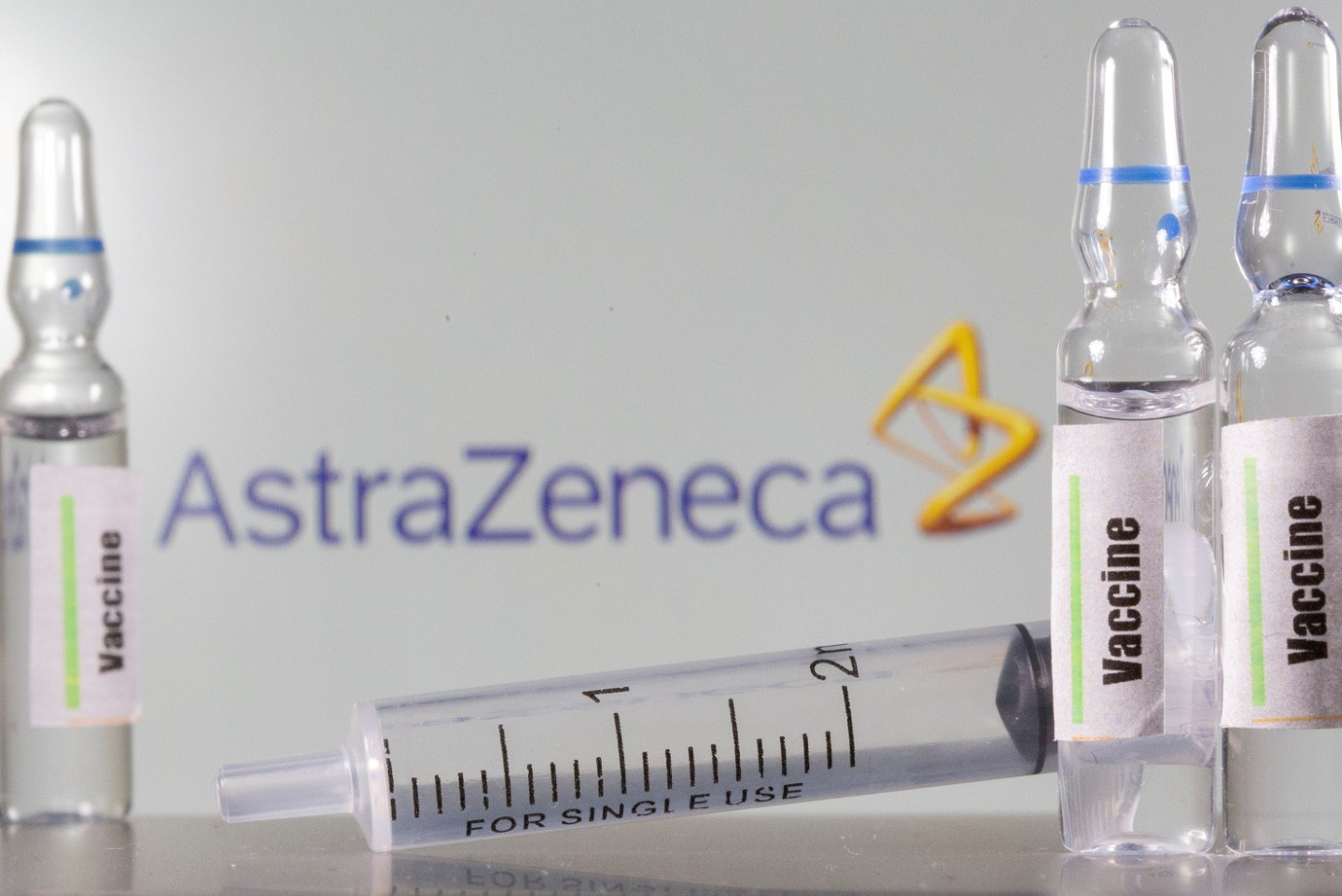 Doses da vacina e uma seringa deitada. Ao fundo, logo da AstraZeneca