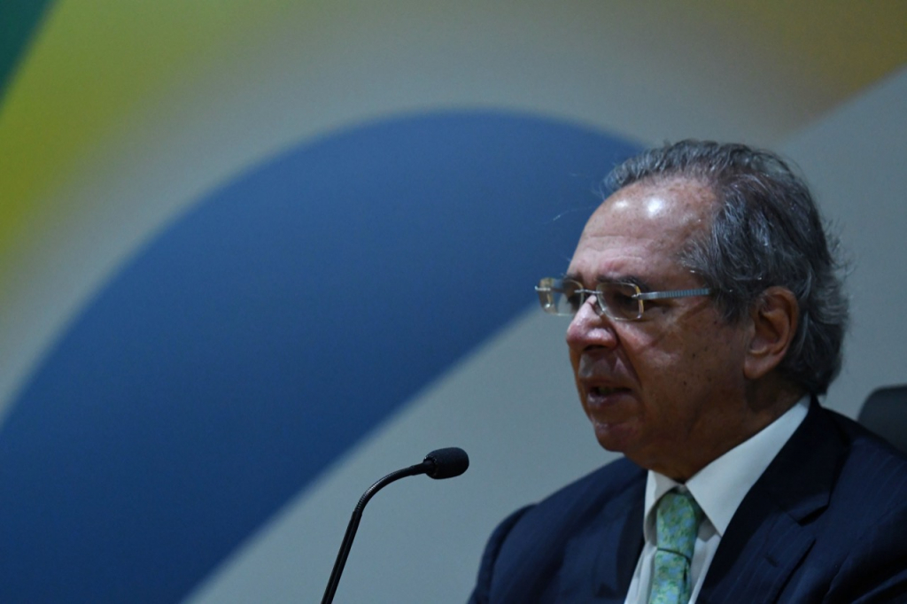 Homem calvo de cabelo grisalho e óculos com armação fina fala em microfone diante da bandeira do Brasil
