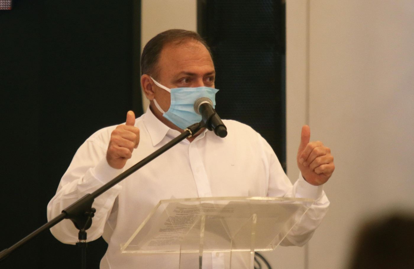 Homem de camisa branca e máscara azul fazendo sinal de positivo com as mãos e falando em microfone