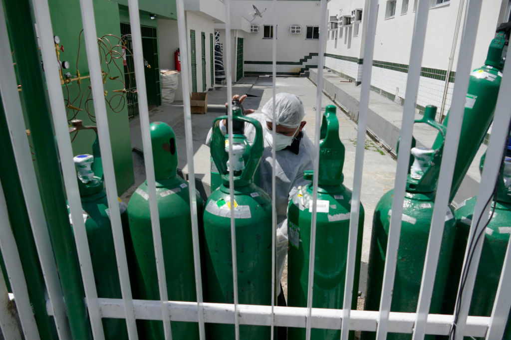 vários cilindros de oxigenio verdes, um sendo manuseado por um profissional da saúde. grade branca em primeiro plano na foto