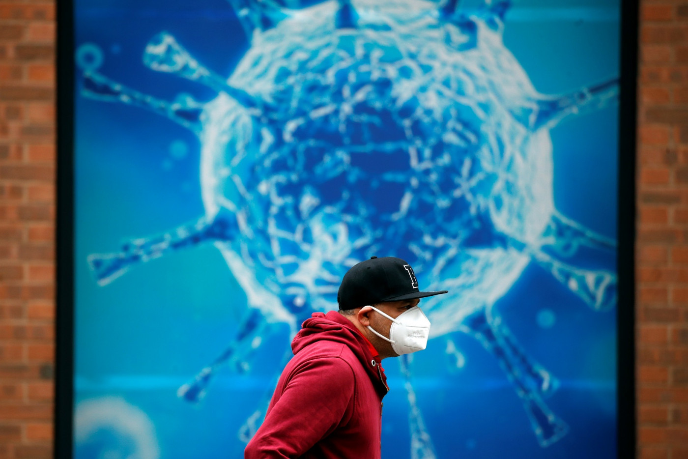 homem andando de máscara com um painel simulando o coronavírus no fundo