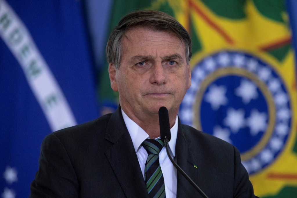 Homem de cabelo grisalho trajando terno e gravata pretos falando em microfone diante de uma bandeira do Brasil
