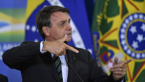 Presidente Jair Bolsonaro de lado, fazendo uma "arminha" com os dedos. Usa terno preto, gravata listrada verde-escura e está na frente de uma bandeira do Brasil.