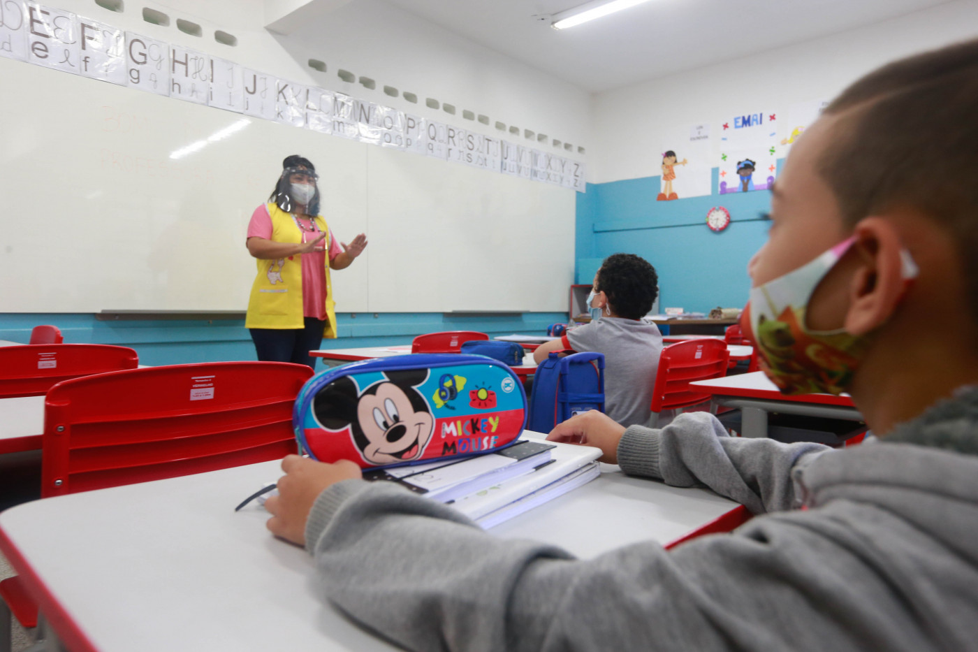 professora usa máscara e face shield enquanto dá aula para crianças em escola