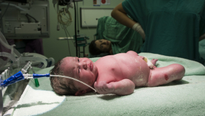 Bebê recém-nascido dentro de hospital; mãe aparece ao fundo após o parto
