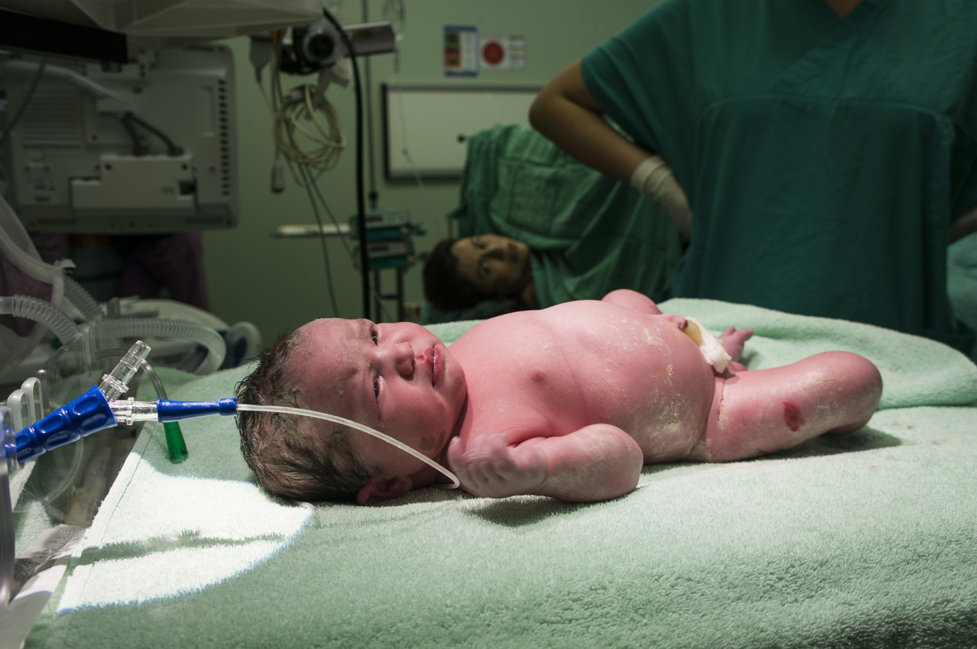 Bebê recém-nascido dentro de hospital; mãe aparece ao fundo após o parto