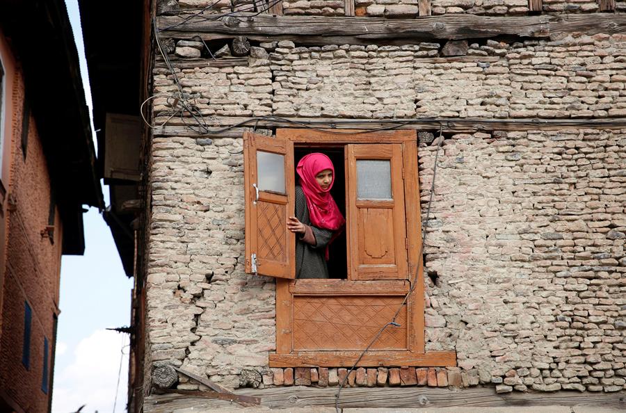 Menina com véu islâmico cor-de-rosa espia pela janela de casa na região da Caxemira, disputada pela Índia e pelo Paquistão