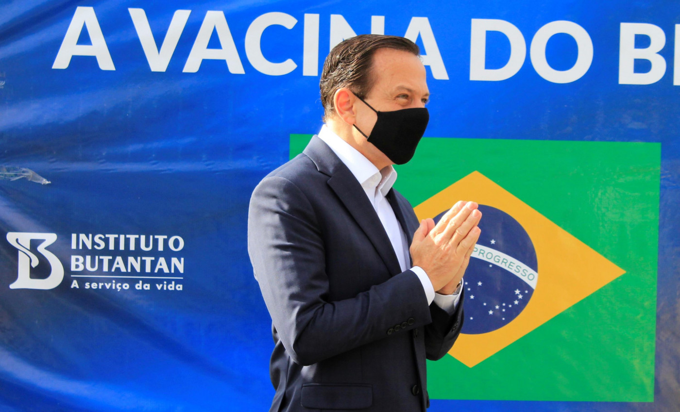 Homem de terno e gravata usando máscara preta e com as mãos unidas diante de uma placa com a bandeira do Brasil, o logo do Instituto Butantan e os dizeres 'a vacina do Brasil'