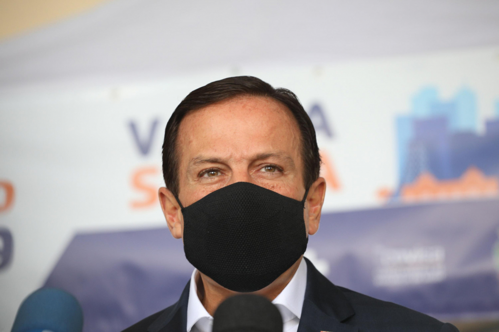 Homem de terno e gravata usando máscara preta diante de fundo com os dizeres 'vacina salva'