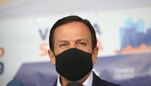 Homem de terno e gravata usando máscara preta diante de fundo com os dizeres 'vacina salva'