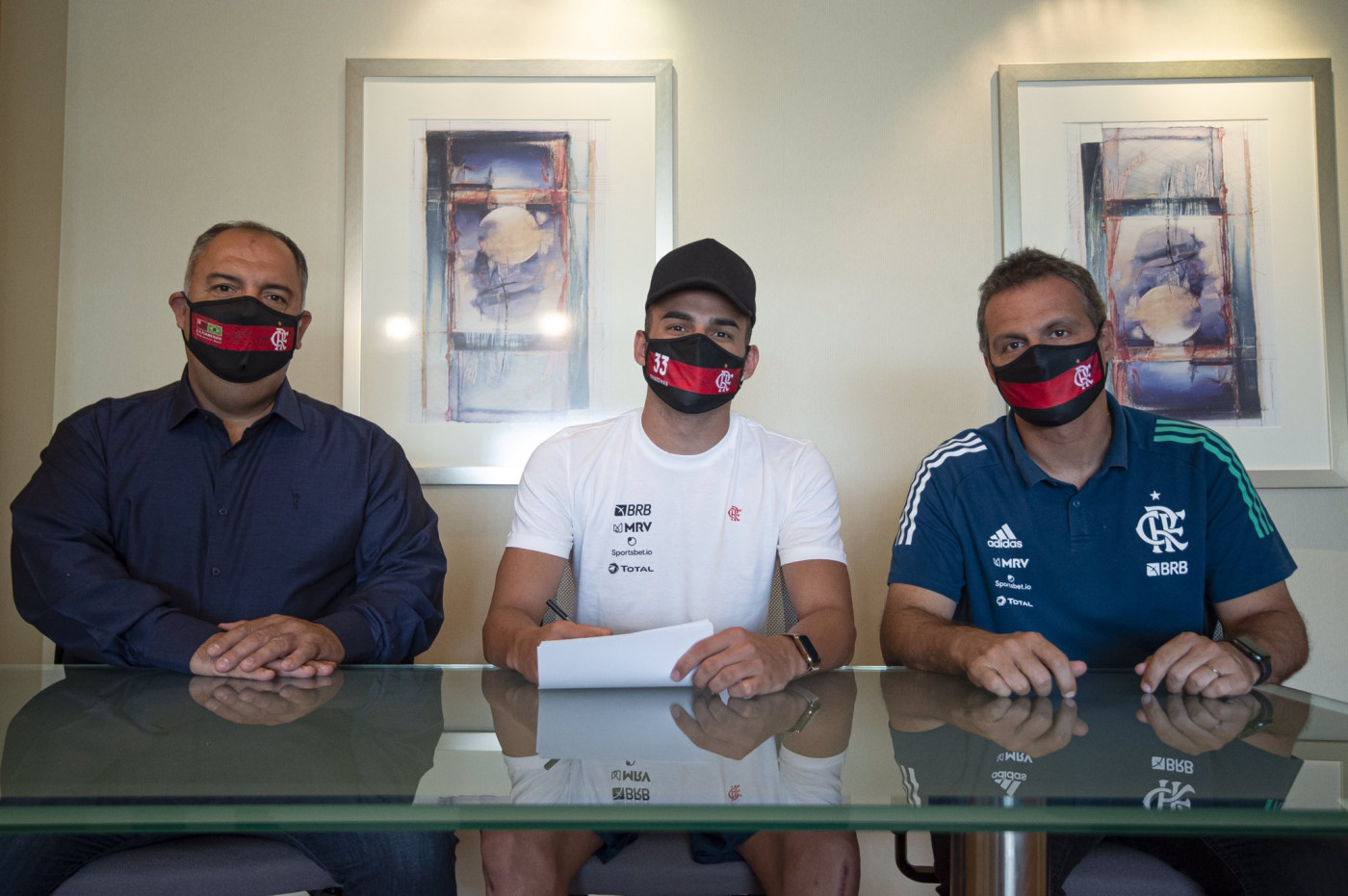 três homens brancos, o do meio Thiago Maia, jogador do flamengo. estão sentados um ao lado do outro e usam máscaras do Flamengo
