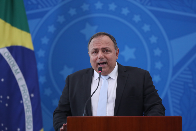 Homem de terno e gravata falando em microfone diante de fundo azul e ao lado de bandeira do Brasil