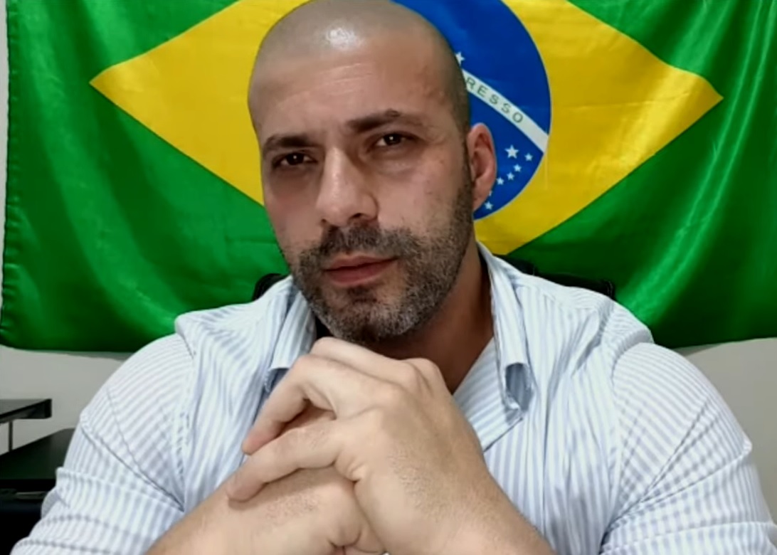 homem careca de blusa azulada com mãos cruzadas diante da bandeira do brasil