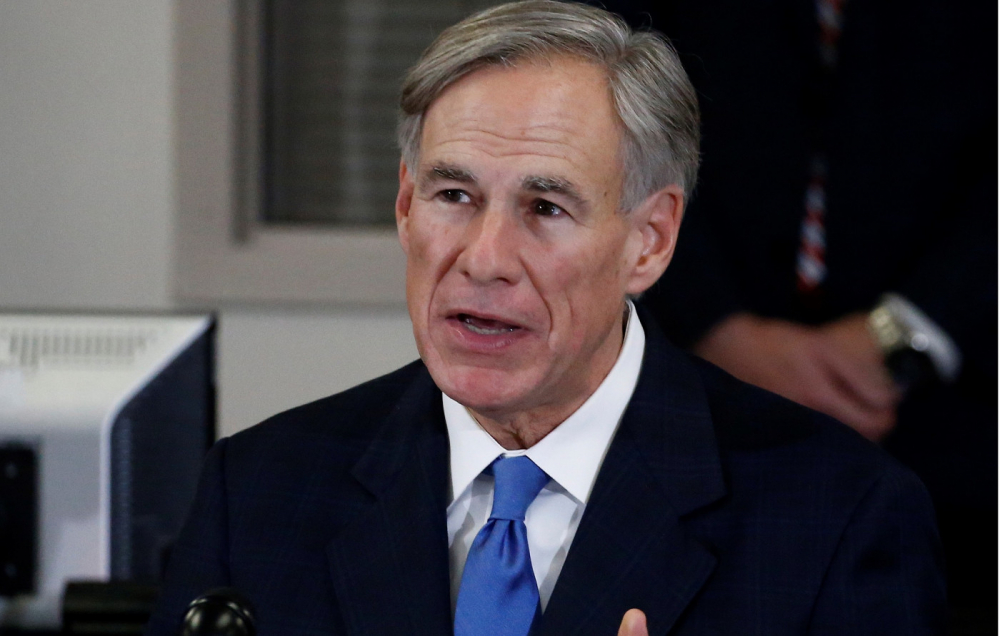 Homem de cabelo grisalho usando terno preto e gravata azul, é possível vê-lo da cintura para cima diante de um fundo desfocado da cor branca