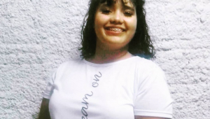 Menina de cabelo preto, aparelho dentário e camisa branca sorri para foto diante de um fundo branco