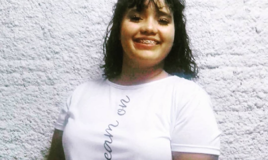 Menina de cabelo preto, aparelho dentário e camisa branca sorri para foto diante de um fundo branco