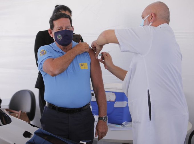Homem de máscara e camisa azul sendo vacinado por enfermeiro careca, de máscara branca e trajes brancos