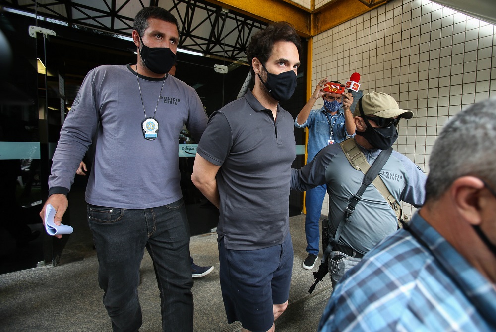 De camisa polo, bermuda e máscara, o vereador Doutor Jairinho é conduzido por policial; ele está algemado, mas suas mãos, posicionadas nas costas, não aparecem na imagem