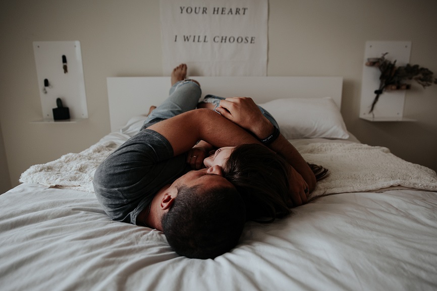 Casal se abraçando deitados em uma cama de lençóis brancos. Só dá para ver o topo das cabeças de onde a câmera está posicionada.
