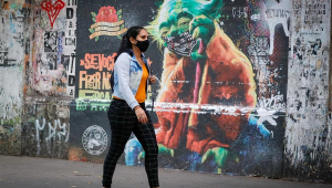 Mulher de máscara caminha em frente a muro grafitado