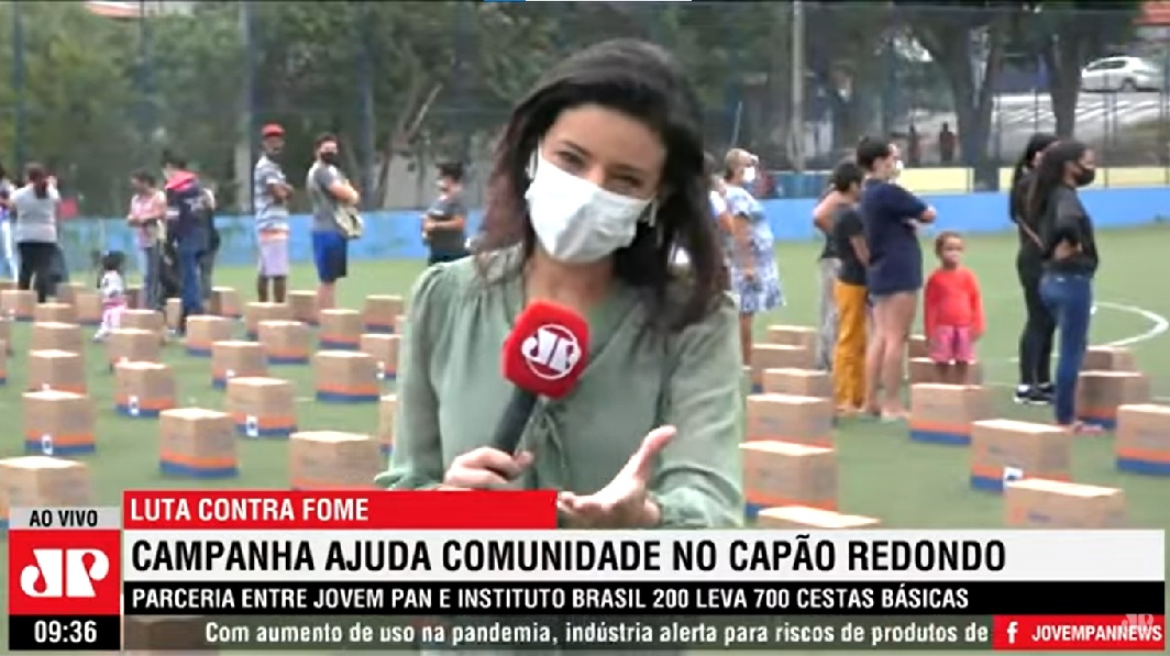 Repórter Carolina Abelin fala do Capão Redondo; no fundo, pessoas fazem fila para receber cesta básica